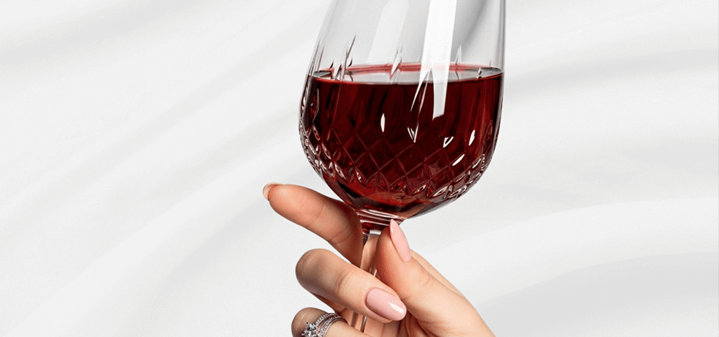 A manicured hand with luxury nails holds a cut-patterned wine glass.
